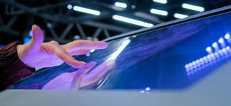 <p>A hand hovering over a public-facing interactive screen</p>
