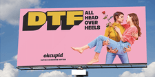 A bright pink billboard showing two women embracing, one holding a long-stemmed red rose in her hand. The heading text says "DTFall Head Over Heels."