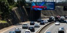 A highway with a digital billboard on the side of a bridge overhead. The billboard shows an ad for a red Lexus.