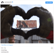 A distant billboard showing Ryan Reynolds and Morena Baccarin smiling and holding hands. In the foreground are two gloved hands making a heart around the distant billboard. The hands are Ryan Reynolds' in costume for Deadpool.