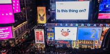 A busy New York City street with various billboards around. One looks like a giant Tweet with the words &quot;Is this thing on?&quot;