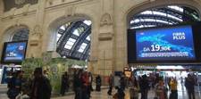 A crowd of people walking through an Italian train station. Overhead is a large digital sign displaying an ad.