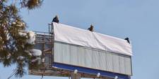 Three workers on top of a billboard, about to unfurl a poster that is due to be posted